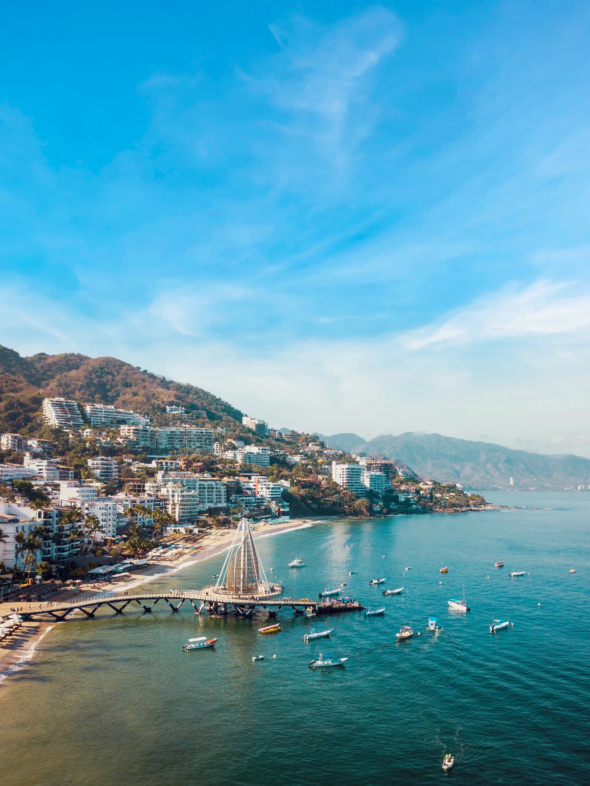 Aerial view of Los Muertos Pier and the Amapas coastline in Puerto Vallarta, featuring hillside condominiums, boats in the blue ocean, and lush coastal mountains.