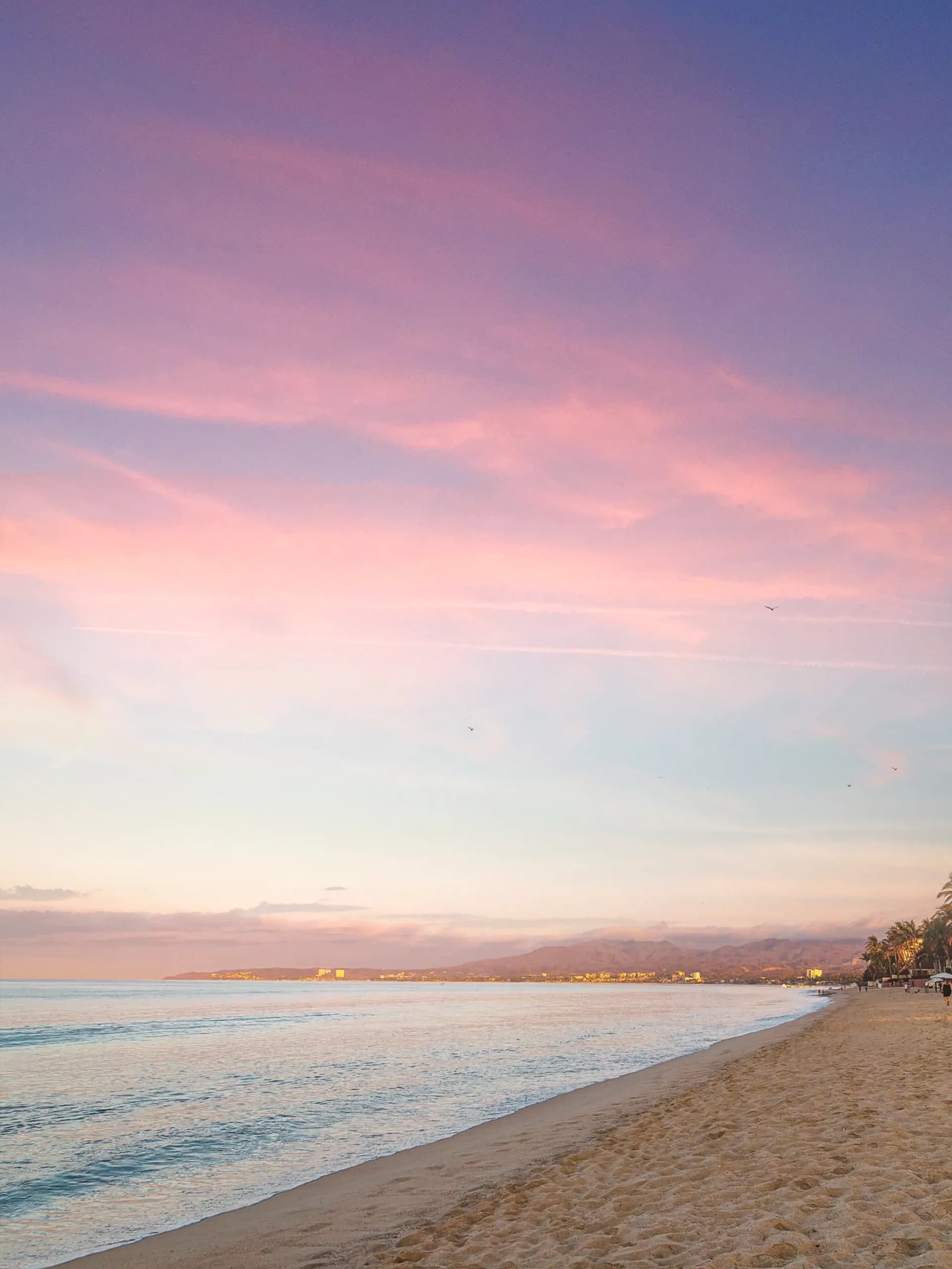 A peaceful sunset stroll along the wide beach of Bucerias, with the sky painted in soft pink and purple hues reflecting over the sand and Banderas Bay.