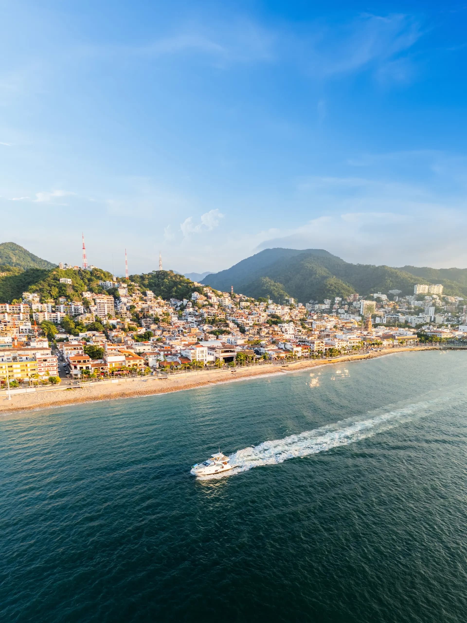 Aerial view of the Puerto Vallarta Malecon area, featuring a white yacht speeding through the blue ocean, a sandy beach, and a bustling coastal town with traditional architecture nestled against lush green mountains.